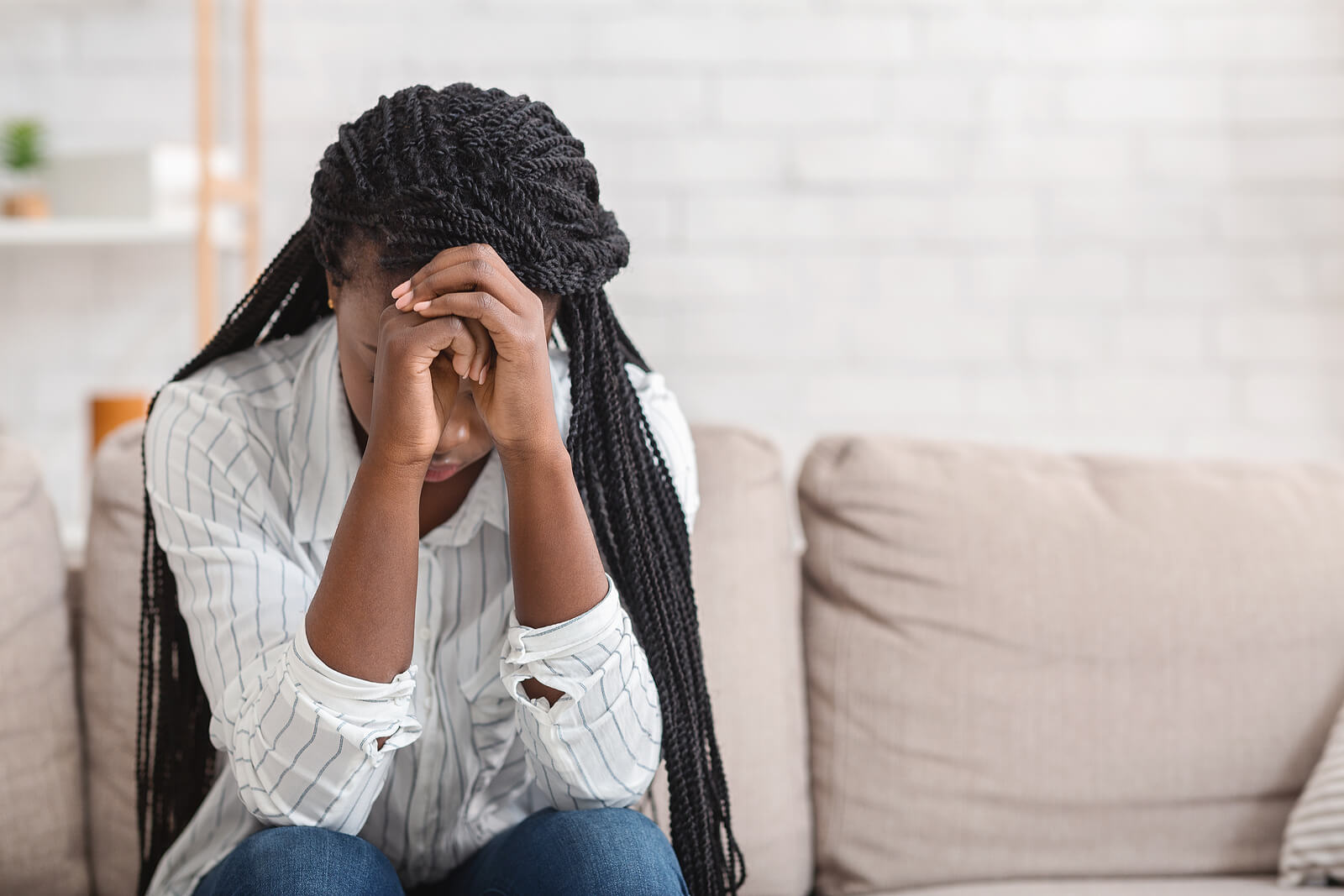 A woman touches her hands to her head while sitting on the couch. This could represent the stress young adults face that black therapists in Brooklyn, NY can address. Learn more about therapy for young adults in Brooklyn, NY by contacting a black therapist in Brooklyn, NY today.