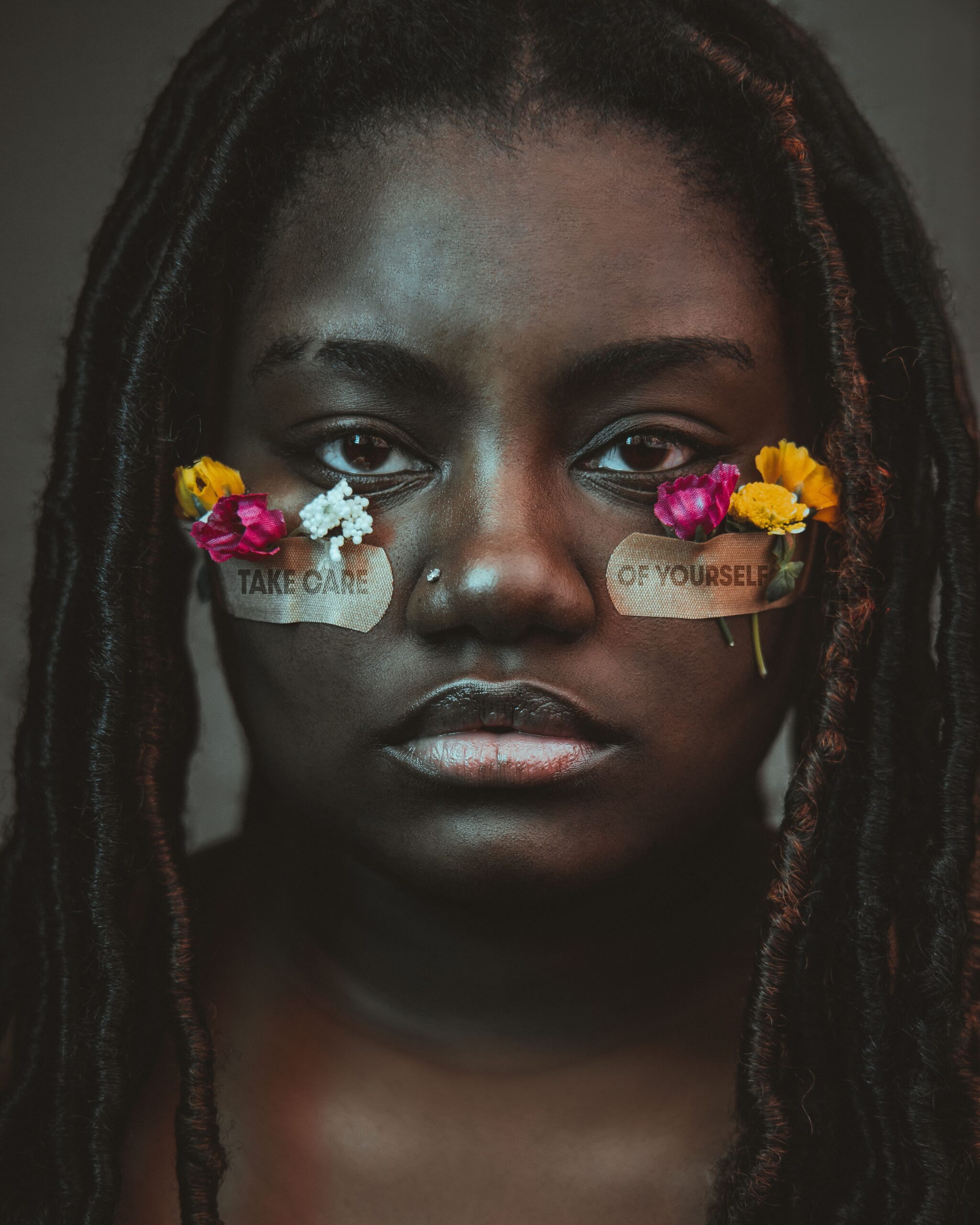 A close up of a black woman with flowers tapped to her cheeks using bandages. The text on the bandages says "take care of yourself". Learn how a black therapist in Brooklyn, NY can offer support with new self care practices. Search for therapy for people of color in Brooklyn, NY to support bipoc young adults today. 