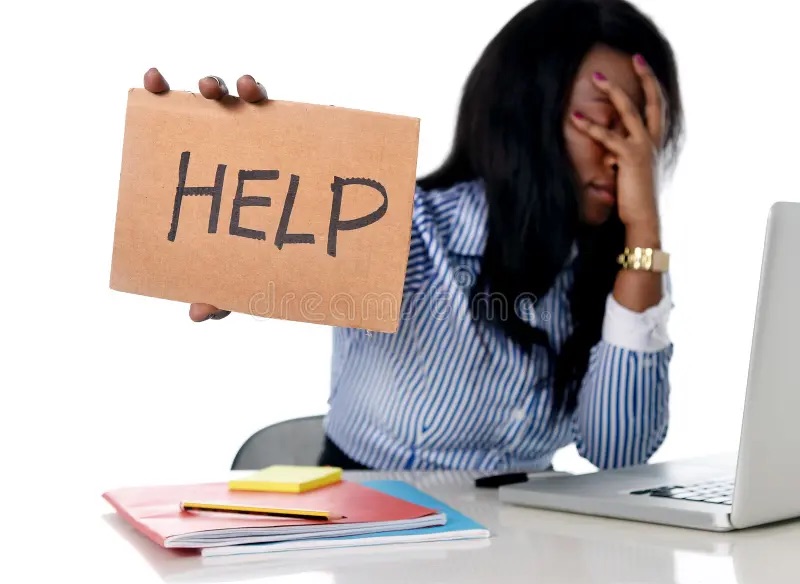 A distressed woman sits at a desk with her head in her hand, holding a cardboard sign that reads “HELP,” symbolizing the urgent need for therapy for anxiety in Brooklyn, NY. This image reflects the emotional overwhelm that can be supported through individual therapy in Brooklyn, NY and working with an attachment style therapist in Brooklyn, NY.