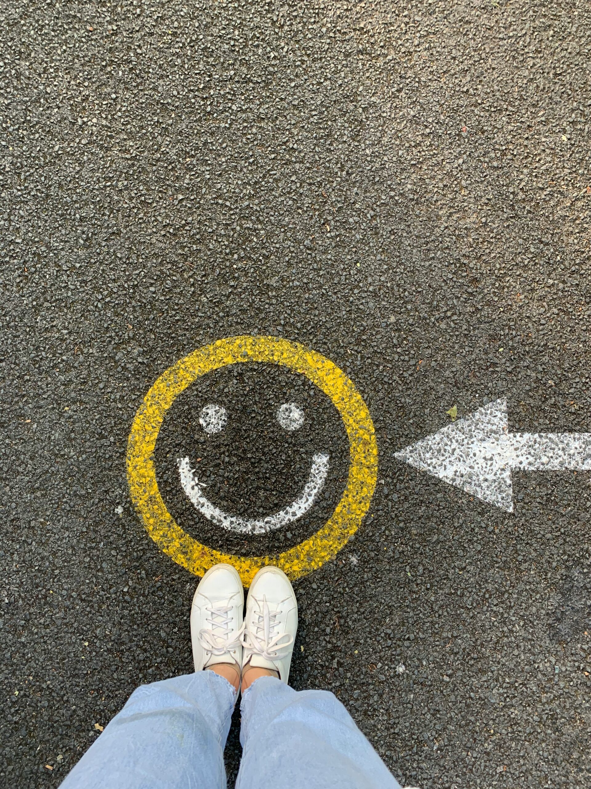 A person stands on pavement where a yellow smiley face and arrow are painted, symbolizing choosing a path toward happiness. Many take this step with anxiety treatment in Brooklyn, NY, guided by a young adult therapist in Brooklyn, NY or through convenient sessions with an online therapist in Brooklyn, NY.