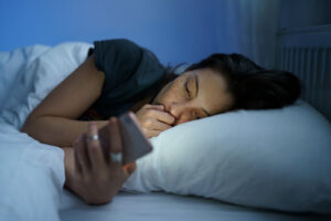 Young woman scrolling on her phone in bed late at night, feeling overwhelmed by options. Gentle counseling for young adults in Brooklyn, NY can ease the spiral. Meet with a therapist for young adults in Brooklyn, NY or choose online therapy in New York for flexible support. 