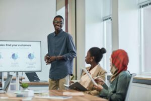 A Black man smiles while presenting to diverse colleagues in a modern office, reflecting the workplace pressures that therapy for anxiety in Brooklyn, NY addresses—work with an anxiety therapist in Brooklyn, NY at SG Wellness.