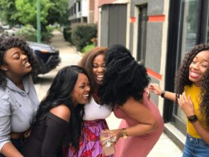 Four young Black women laugh and hug on a city sidewalk, reflecting the joy and connection that anxiety treatment in Brooklyn, NY helps restore—connect with a therapist for young adults in Brooklyn, NY at SG Wellness.