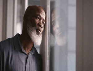 An older Black man with a white beard gazes out a window with a somber, reflective expression, capturing the weight of world anxiety that anxiety treatment in Brooklyn, NY addresses — a culturally sensitive therapist in Brooklyn, NY at SG Wellness can help you find peace.
