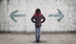 A young woman with red hair stands before a concrete wall painted with arrows pointing in opposite directions, capturing the disorienting feeling of being stuck in a loop when relationships and values begin to pull in different directions. For young adults navigating this kind of uncertainty, working with a young adult therapist in Brooklyn, NY can help bring clarity to what once felt like an impossible choice. 