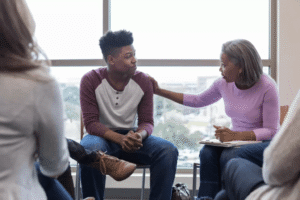 A therapist places a supportive hand on a young person's shoulder during what appears to be a group therapy session, representing the compassionate, personalized support that therapy for self-esteem in Brooklyn, NY can provide. Building self-esteem in Brooklyn, NY often begins with feeling truly seen and supported in a safe therapeutic environment. A self-esteem therapist in Brooklyn, NY can help you understand the roots of negative self-beliefs and develop healthier, more confident ways of relating to yourself and others. 