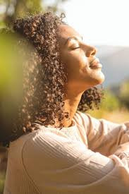 A woman with curly hair tilts her face toward warm, golden sunlight with her eyes closed, embodying the relief and openness that becomes possible when you stop being stuck in a loop and begin to move through difficult emotions. This kind of peace is what counseling for young adults in Brooklyn, NY works toward — helping you feel grounded, present, and reconnected with yourself. 