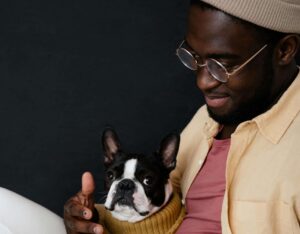 A Black man wearing glasses and a beanie smiles while sitting on a couch with his Boston Terrier and a laptop, representing the comfort and accessibility of working with an anxiety therapist in Brooklyn, NY through therapy for anxiety in Brooklyn, NY at SG Wellness.
