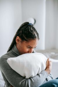 A young woman of color hugs a pillow with a pained expression, reflecting the isolation of anxiety—a young adult therapist in Brooklyn, NY and anxiety therapist in Brooklyn, NY at SG Wellness can help.