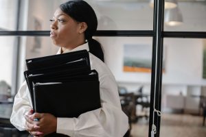 A professional woman in a white blouse holds a stack of heavy binders, gazing off with a serious, thoughtful expression. The weight of having to prove yourself is real. A self-esteem therapist in Brooklyn, NY, self confidence therapy in the Bronx, NY, and a self esteem therapist in Queens, NY help lighten that load.