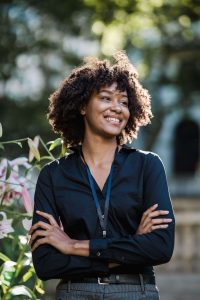 A confident professional stands with her arms crossed and a relaxed smile, wearing a work lanyard outdoors. This is what internal confidence can look like. Improving self-esteem in Brooklyn, NY with a self-esteem therapist in Brooklyn, NY, or a self esteem therapist in Queens, NY, helps success finally feel earned. 