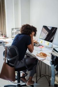 A professional sits at a busy desk with her hands pressed to her head, looking overwhelmed by the pressure to prove herself. Overworking often masks self-doubt. Self confidence therapy in the Bronx, NY, therapy for self-esteem in Brooklyn, NY, and self esteem therapy in Queens, NY can interrupt that cycle. 